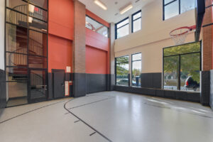 Interior basketball court, half court, spiral staircase, padded walls, gray floor, red wall, large windows.