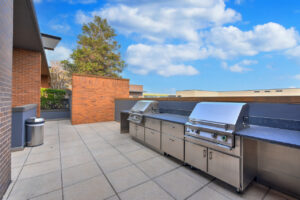 Outdoor Rooftop Grill Area, 2 large grills, red brick walls, bushes and tree in background, photo taken on a sunny day.