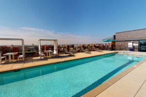 Exterior Rooftop Pool area, covered dining tables, lawn chairs, skyline in the background at sunset.